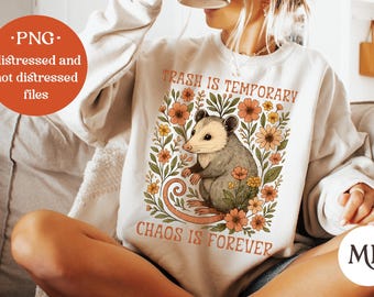 May include: A cream-coloured sweatshirt featuring the text "Trash is Temporary, Chaos is Forever" and an opossum graphic surrounded by flowers. The design incorporates orange, green, and cream hues. The image also includes a PNG file label.