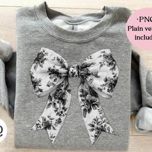 Puede incluir: Una sudadera gris con un diseño de lazo grande. El lazo es blanco con un estampado floral negro. El texto "PNG Plain version included" está en un círculo rosa. La sudadera tiene cuello redondo y mangas largas.