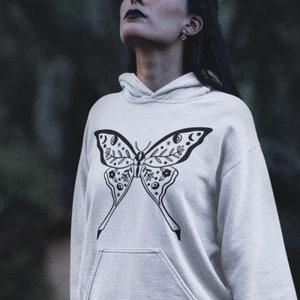 Może przedstawiać: Biała bluza z kapturem z czarno-białym wzorem motyla. Motyl ma skomplikowane detale, w tym elementy kwiatowe i symbole niebieskie.