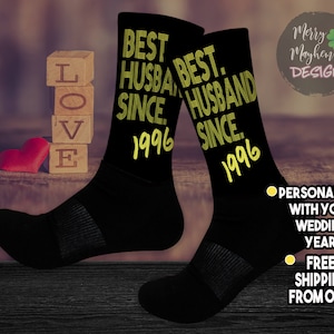 May include: A pair of black crew socks featuring yellow text that reads BEST HUSBAND SINCE 1996. The design includes a ribbed cuff and a reinforced heel and toe area. The text is printed in a bold, sans-serif font with a stacked layout on the leg.