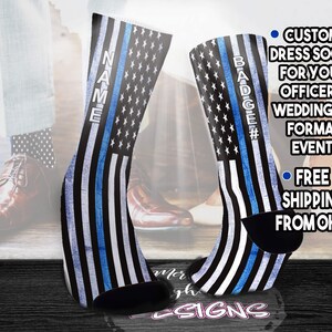 May include: A pair of black dress socks featuring a thin blue line and a white star pattern on a dark background. One sock has the word NAME printed vertically, and the other has BADGE# printed vertically. The socks are displayed against a blurred background.