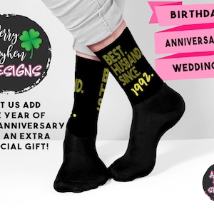 May include: A pair of black crew socks featuring the yellow text "BEST. HUSBAND. SINCE. 1992." printed on the side. The socks are worn on the feet, which are positioned against a white background with pink banners labeled "BIRTHDAY", "ANNIVERSARY", and "WEDDING".