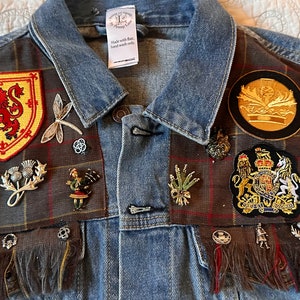 May include: A blue denim jacket with a plaid fabric panel on the front. The panel is covered in various patches, pins, and charms, including a Scottish lion, a thistle, a dragonfly, and a crown. The text "OUTLANDER The Series" is visible on a black patch.