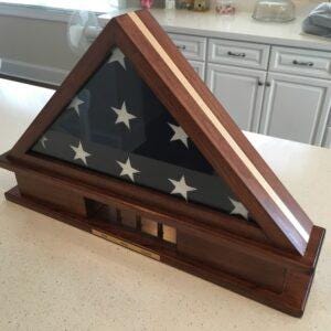 May include: A triangular wooden display case with a dark brown finish and a light wood inlay stripe. The case features a glass front panel protecting a folded American flag with visible white stars on a dark blue field. A small gold plaque is attached.