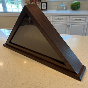May include: A dark brown wooden triangular shadow box with a glass front. The shadow box is designed to display a folded American flag.
