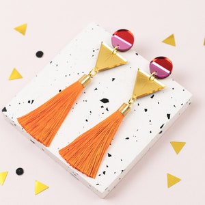 Puede incluir: Un par de pendientes de borla naranja con detalles de triángulo dorado y círculo rosa y rojo.