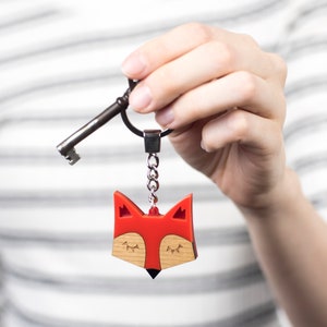 May include: A red fox keychain with closed eyes and a wooden detail. The keychain is attached to a silver chain and a key.