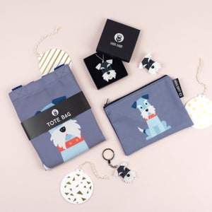 Könnte beinhalten: Ein Set aus grauer und blauer Tragetasche, Beutel, Schlüsselanhänger und Ornamenten mit einem Schnauzer-Hundemuster. Die Tragetasche hat den Text "TOTE BAG" aufgedruckt. Der Beutel hat den Text "LAURA DARBY" aufgedruckt. Der Schlüsselanhänger hat einen Schnauzer-Hund-Anhänger. Die Ornamente sind rund und haben einen weißen Hintergrund mit goldenen Dreiecken.