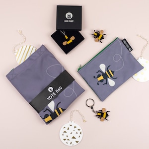May include: A set of bee-themed tote bags, pouches, and keychains. The items are all a light grey colour with a black and yellow bee design. The tote bag has the text "TOTE BAG" printed on it. The pouches have a zipper closure. The keychains are made of metal and have a bee charm.