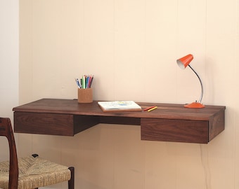Op de afbeelding: Een aan de muur gemonteerd zwevend bureau van donker hout met twee lades, een cilindrische pennenhouder met kleurpotloden, een notitieboek en een oranje bureaulamp. Een houten stoel met een gevlochten zitting staat voor het bureau.