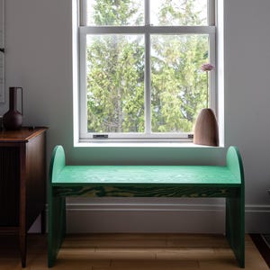 May include: A bright green wooden bench with curved backrests sits beneath a window. The window frames a view of tall green trees. A brown vase and a dark brown cabinet are also visible.