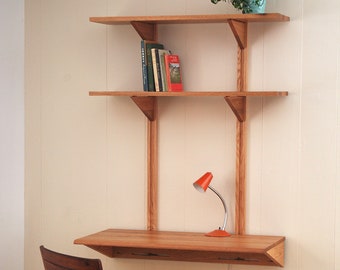May include: A wooden wall-mounted desk with a shelf above it. The desk has a light wood finish and a simple design. There is a small lamp on the desk and a chair in front of it.
