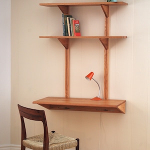 Puede incluir: Un escritorio de madera montado en la pared con dos estantes superiores y una superficie de trabajo inferior. La unidad incluye una pequeña lámpara de escritorio naranja, varios libros y una planta en maceta. Una silla de madera con asiento trenzado.