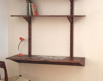Op de afbeelding: Een aan de muur gemonteerd bureau van donker hout met twee bovenste planken ondersteund door verticale beugels. Het bureaublad bevat een kleine oranje lamp en een puzzel. Een houten stoel met een gevlochten zitting staat voor het bureau.