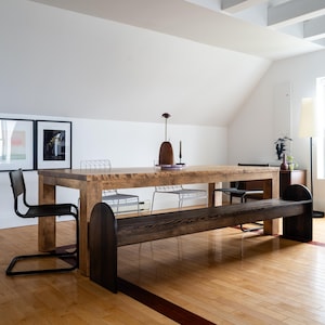 Könnte beinhalten: Ein großer, rechteckiger Esstisch aus Holz mit natürlicher Oberfläche, kombiniert mit einer dunklen Holzbank und Metallstühlen. Der Raum verfügt über Holzböden, weiße Wände und dekorative Elemente, die ein modernes Interieur schaffen.