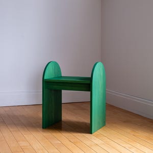 May include: A green wooden stool with a simple, modern design. The stool has a rectangular seat and two arched supports on each side. The wood grain is visible, and the stool is painted in a vibrant green color.