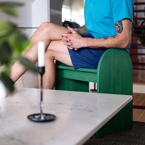 May include: A green wooden bench with a curved back and a white marble coffee table. A black candle holder with a white candle is on the table. The person is wearing a blue t-shirt and blue shorts.