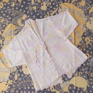 Könnte beinhalten: Ein weißer, leichter Cardigan mit zarter Spitzenborte und einem Bindeverschluss. Der Cardigan hat ein Batik-Muster in Rosa- und Gelbtönen.