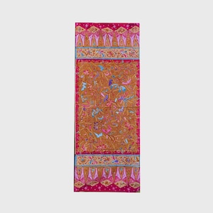 Pode incluir: Um têxtil retangular com um painel central de tecido castanho-dourado com um padrão intrincado de flores e pássaros em tons de rosa, azul e branco. A peça é emoldurada por largas bordas magenta vibrantes com motivos geométricos e florais.