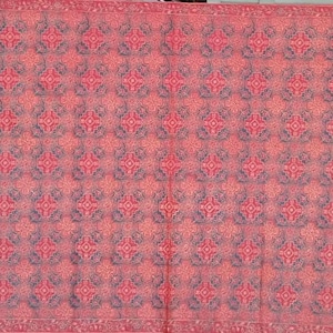 Può includere: Un tessuto rettangolare con un motivo ripetitivo di disegni floreali e geometrici blu scuro su uno sfondo rosso vivace. Il tessuto è incorniciato da bordi verticali contenenti una serie di triangoli blu scuro con dettagli floreali.