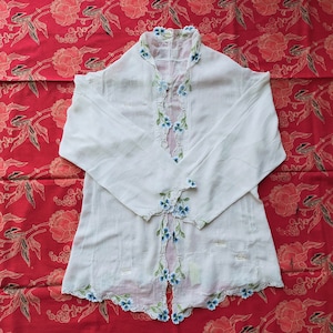 Könnte beinhalten: Eine weiße Baumwollbluse mit zarter blauer Blumen Stickerei. Die Bluse hat einen runden Ausschnitt und lange Ärmel mit einem gewellten Saum. Der Stoff ist durchsichtig und leicht.