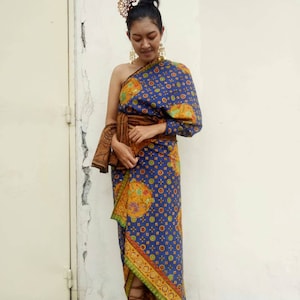 Könnte beinhalten: Eine Frau trägt ein blaues und goldenes gemustertes Kleid mit einem braunen Gürtel. Das Kleid ist einseitig und hat einen langen, fließenden Rock. Sie trägt braune Sandalen und hat einen goldenen Kopfschmuck.