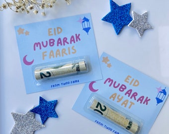 May include: Two blue and silver glitter stars, a white string of pearls, and two blue and white cards with rolled up dollar bills inside. The cards have the text "Eid Mubarak Faaris" and "Eid Mubarak Ayat" with the words "From: Tinoo Faiba" at the bottom.