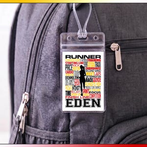 May include: A rectangular clear plastic luggage tag with a white insert featuring a black silhouette of a runner surrounded by red, black, and yellow text including words like RUNNER, PACE, ENDURANCE, and the name EDEN in large black letters.