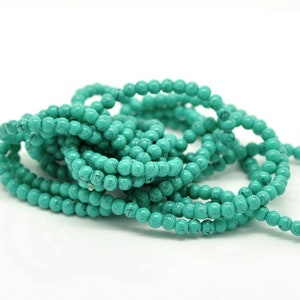 May include: Strands of turquoise-colored beads with a speckled pattern. The beads are small and round, and they are arranged in a tangled, layered formation against a white background. The beads are a vibrant, eye-catching color.