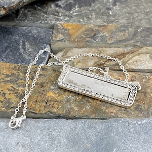 May include: A silver-toned necklace with a rectangular pendant. The pendant is bordered with small, clear crystals and hangs from a delicate chain. The necklace is displayed on a textured, gray and brown stone surface.