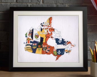 May include: A framed cross-stitch embroidery depicting the map of Canada, with each province and territory filled with its respective provincial or territorial flag design, presented in a black frame on a white background.