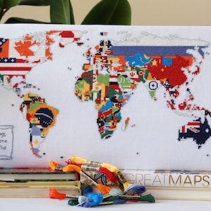 Può includere: Una tela bianca con un ricamo a punto croce di una mappa del mondo con bandiere di diversi paesi. Il testo "Flags of the world" è scritto su un rotolo bianco nell'angolo in basso a sinistra dell'immagine. La tela è incorniciata e appoggiata su una pila di libri con il titolo "GREATMAPS" visibile sul libro superiore.