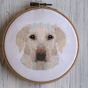 May include: Cross-stitch embroidery of a dog's face in a wooden embroidery hoop. The dog is white with brown markings and has a light brown nose.