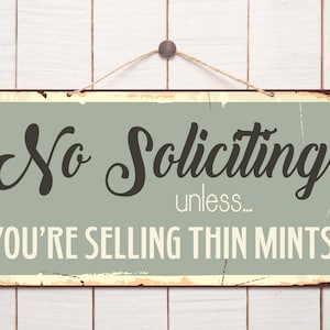 May include: A rectangular metal sign with a distressed finish, featuring the words "No Soliciting" in a bold, vintage font. Below, it reads "unless... YOU'RE SELLING THIN MINTS". The sign is mounted on a white wooden background.