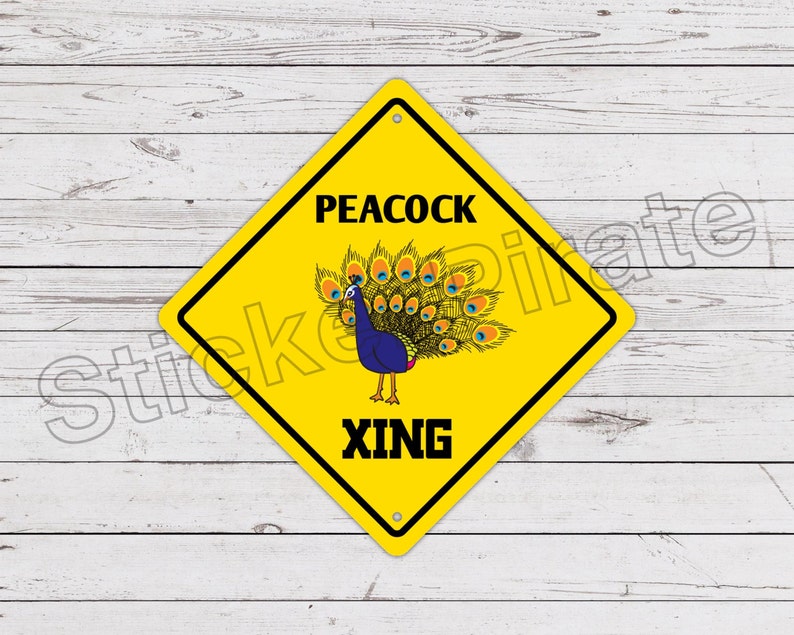 Op de afbeelding: Geel ruitvormig bord met zwarte rand en tekst "PEACOCK XING". Het bord toont een blauwe pauw met een kleurrijke staart. De achtergrond is een wit houten oppervlak.
