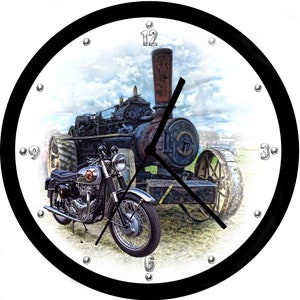 May include: A vintage clock with a black frame and white face. The clock features a black and red steam engine and a silver motorcycle on a grassy field. The clock face has silver Roman numeral hour markers and silver hands.
