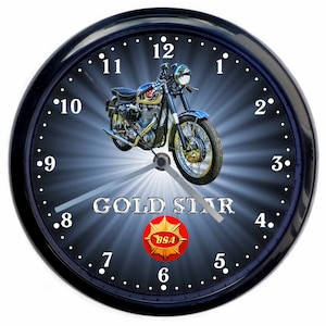 May include: A black wall clock with a silver face and black hands. The clock face features a vintage motorcycle with the words "GOLD STAR" and a red and gold BSA logo. The clock has a black frame with white numbers.
