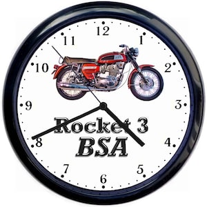 Op de afbeelding: Een klok met een zwart frame en een witte wijzerplaat met een rode en chromen motorfiets met de tekst "Rocket 3 BSA" eronder. De wijzerplaat van de klok heeft zwarte cijfers en wijzers.