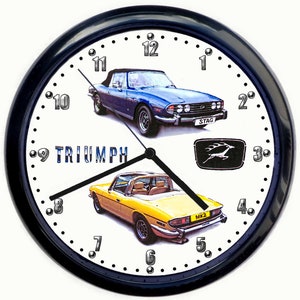 May include: A black wall clock with a white face featuring two Triumph Stag convertibles. The clock has silver numbers and a black hour and minute hand. The top car is blue and the bottom car is yellow. The clock face also includes the word "TRIUMPH" and the Triumph Stag logo.