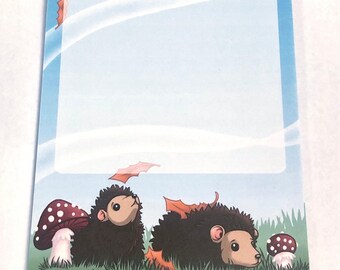 May include: A blue notepad with a white border and a white background. The notepad has a design of two hedgehogs with brown fur and pink noses. The hedgehogs are surrounded by green grass and brown leaves. The text "www.msmorrillillustration.com" is printed at the top of the notepad.