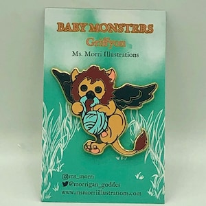 May include: A gold-colored enamel pin featuring a cartoon baby griffin with brown fur, blue eyes, and a yellow ball of yarn. The griffin has wings and a lion tail. The pin is on a green and white background with the text "Baby Monsters Griffon Ms. Morri Illustrations" and social media handles.