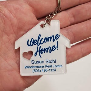 May include: A clear, house-shaped keychain with the words "welcome Home!" in blue script. A heart-shaped cutout is visible. The keychain includes the name "Susan Stohl" and contact information for Windermere Real Estate.