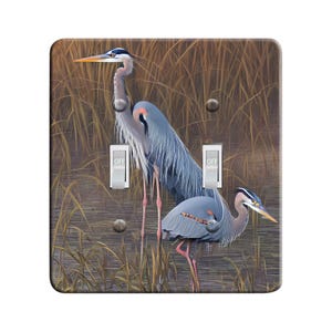 May include: A light switch cover featuring a realistic painting of two blue herons standing in a marsh setting. The herons are gray and blue with long legs and necks. The background is a brown and green marsh with tall reeds.