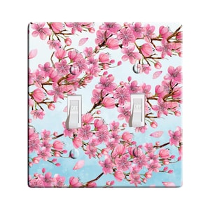 May include: A light blue and white light switch cover with a pink cherry blossom floral pattern. The switch cover has two toggle switches labeled "OFF".