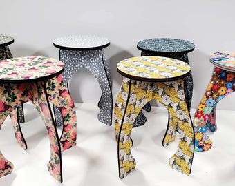 May include: A collection of six decorative stools with floral and patterned designs. Each stool has a round top and curved legs. The color palette includes pink, yellow, blue, and green, with black outlines. The stools are made of wood.