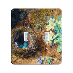 May include: A decorative light switch cover featuring a detailed painting of a bird's nest with blue eggs, yellow flowers, and the word "OFF" on the switches. The artwork has a natural, earthy color palette.