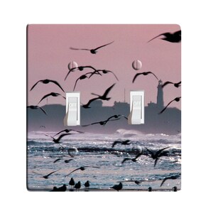 May include: A light switch plate with a scenic image of a lighthouse and seagulls flying over a body of water. The switch plate has two toggle switches labeled "OFF".
