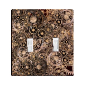 May include: A decorative double light switch cover featuring a steampunk design. The cover is covered with a detailed pattern of gears in shades of brown, gold, and bronze. Two white switches are in the off position.
