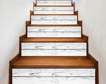 May include: Wooden staircase with white, distressed wood-look stair treads. The treads have a weathered appearance, with visible wood grain and gray accents. The risers and side panels are a warm, medium brown. The stairs lead upwards against a white wall.