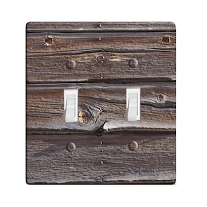 May include: A decorative light switch cover featuring a weathered wood design. The cover has two white toggle switches, both in the "OFF" position. The wood grain is visible, with knots and nail heads adding texture.